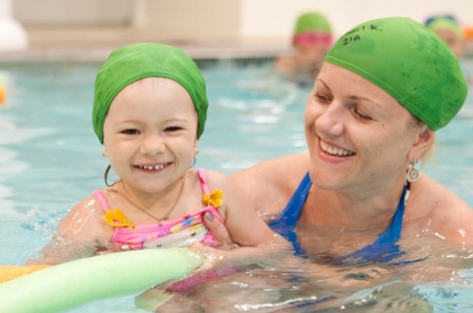Mom holding daughter in pool with green caps and green noodle.