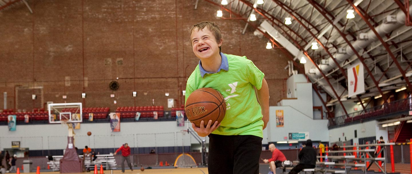 Boy in green t-shirt smiling with basketball at indoor YMCA gymnasium