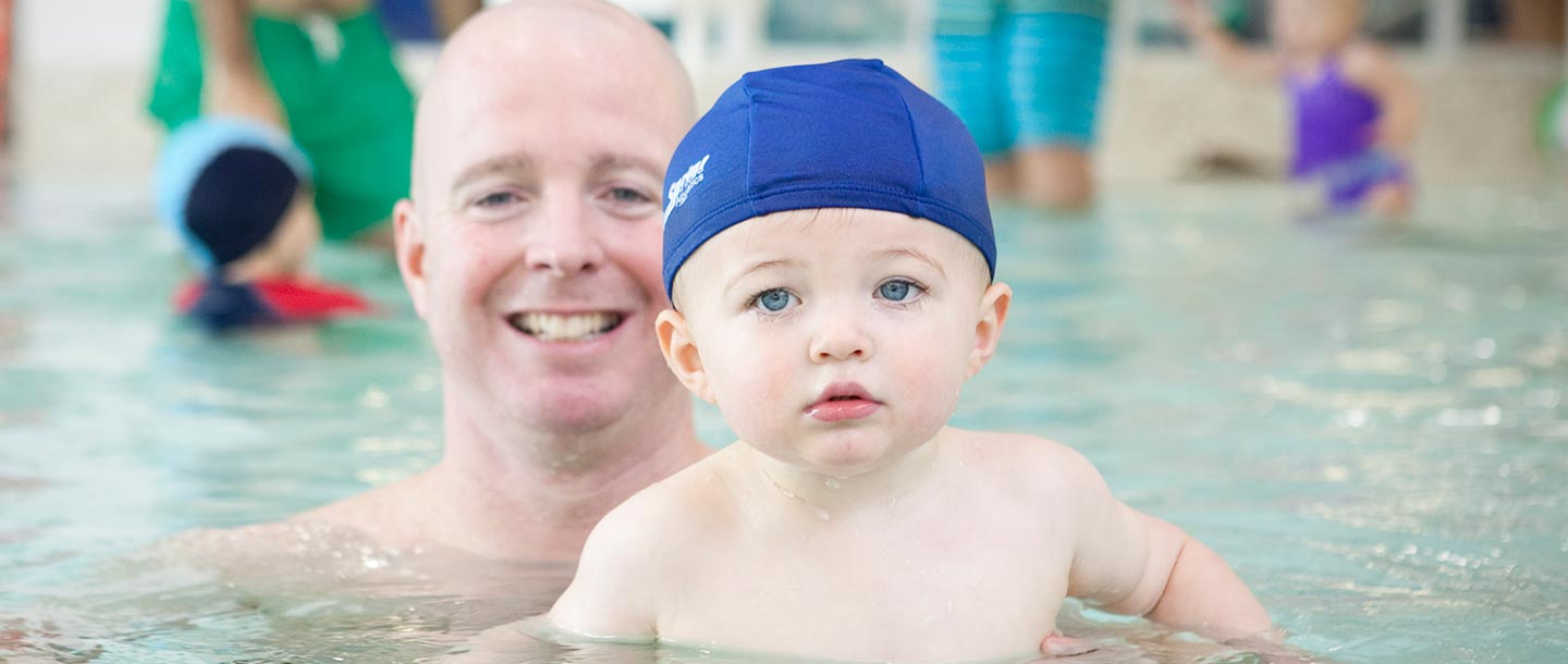 Dad with baby in blue swim cap swimming at YMCA indoor pool