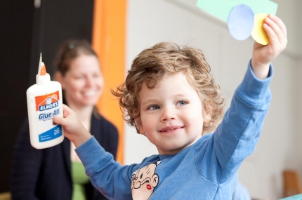 Toddler holding glue and art project with arms up at the Prospect Park YMCA class