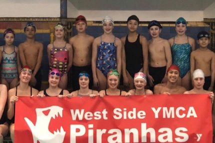 West Side YMCA Piranhas swim team sitting on side of indoor pool holding swim team sign