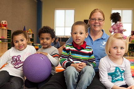 YMCA staff member sitting on floor of child playroom with four kids and purple ball