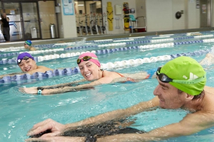 Three adults swimming in Vanderbilt YMCA lap pool with kickboards and smiling at each other