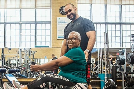 Woman wearing glasses smiling on weight machine at West Side YMCA strength training room with smiling personal trainer