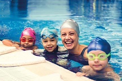 Woman and three kids wearing caps and googles smiling and swimming in pool