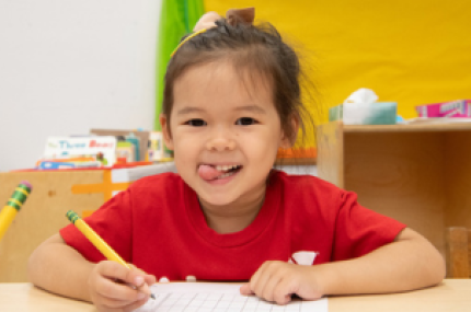 Young girl in classroom smiling at the camera while working on an assignment.