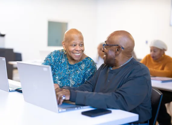 An older Black couple smiles at each other during a computer literacy class.