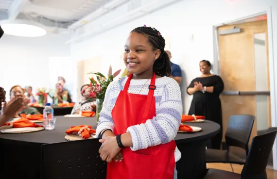 Kid in apron serves food to dinner guests at NEBX Teaching Kitchen culminating event.