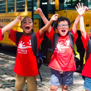 Four kids with arms up on YMCA field trip during summer camp in front of school bus