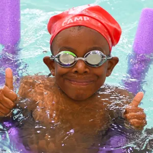 Summer camp boy in indoor pool with goggles, noodles, and thumbs up.