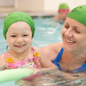 Mom holding daughter in pool with green caps and green noodle.