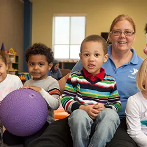 YMCA staff member sitting on floor of child playroom with four kids and purple ball