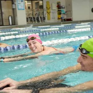 Three adults swimming in Vanderbilt YMCA lap pool with kickboards and smiling at each other