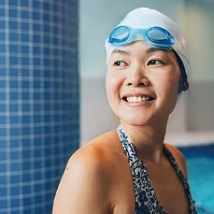 Woman with blue googles and swim cap smiles in indoor swimming pool