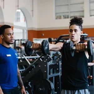 Fitness coach watches teen lifting dumbbells in YMCA weight lifting room at Ridgewood YMCA