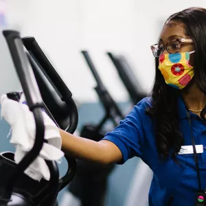 YMCA staff member in mask cleans exercise equipment.