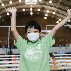 A boy in a mask smiling and waving during camp at the YMCA.