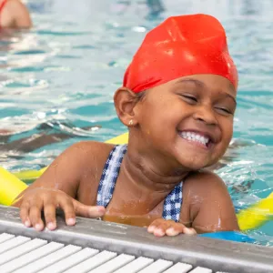 young child smiling and hanging onto the side of the pool with a yellow pool noodle