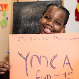 smiling grade-school student holding sign that says YMCA equals FUN