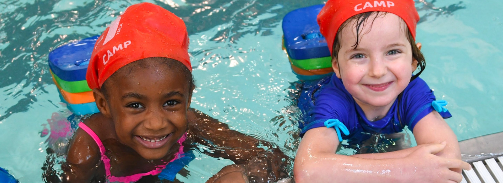 Two kids in YMCA swim caps smile from the pool at the Dodge YMCA summer day camp in Brooklyn.