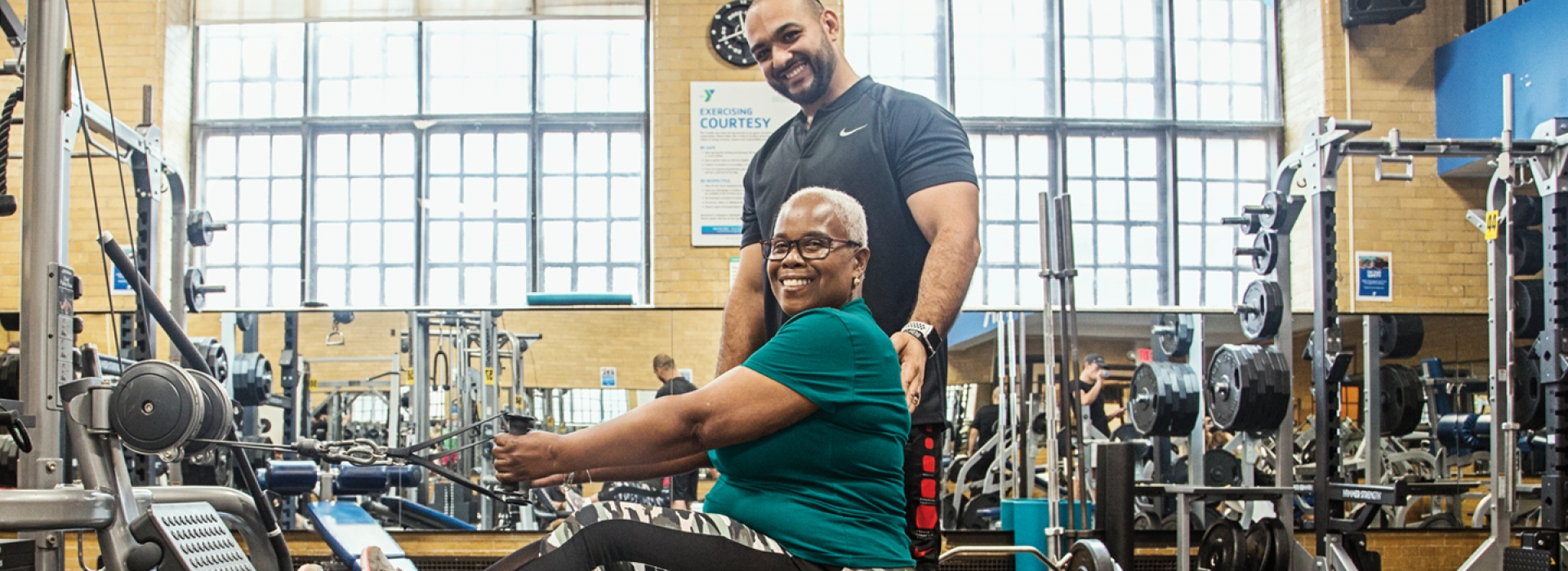 A woman works on strength training machine with a personal trainer at the YMCA.