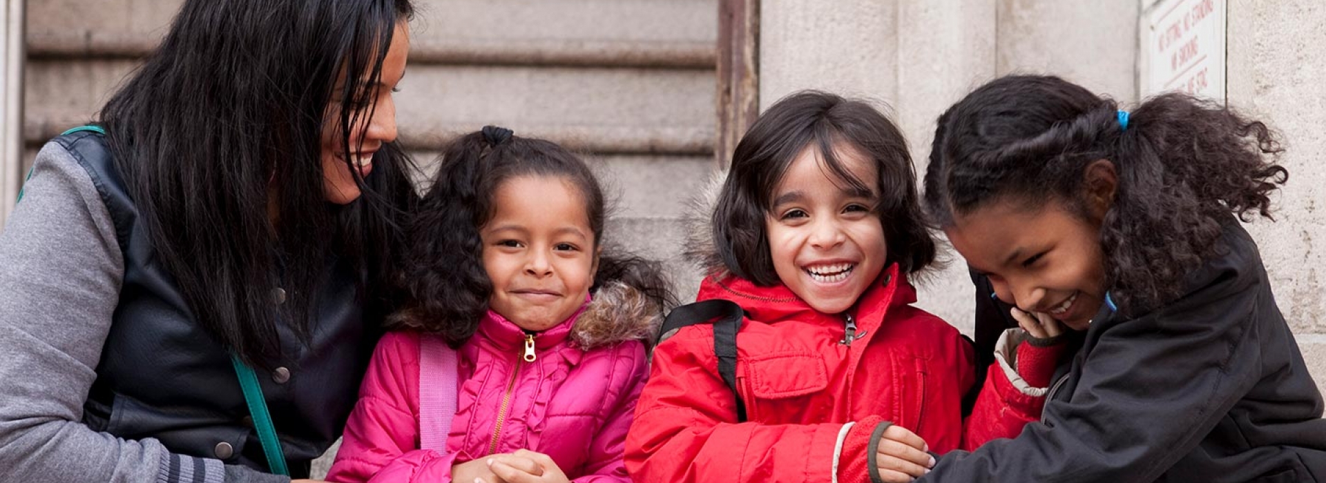 Mom and three kids in winter coats sit outside on steps of Greenpoint YMCA in Brooklyn