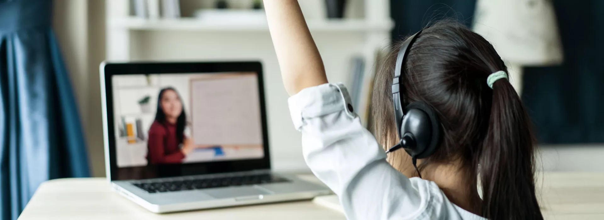 A child in headphones looks at an instructor on a laptop and raises her hand.