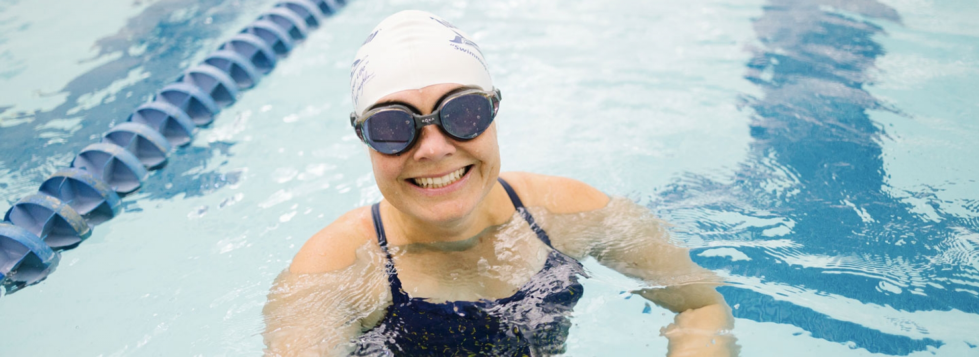 A woman in a swim cap and goggles smiles in the pool at the YMCA.