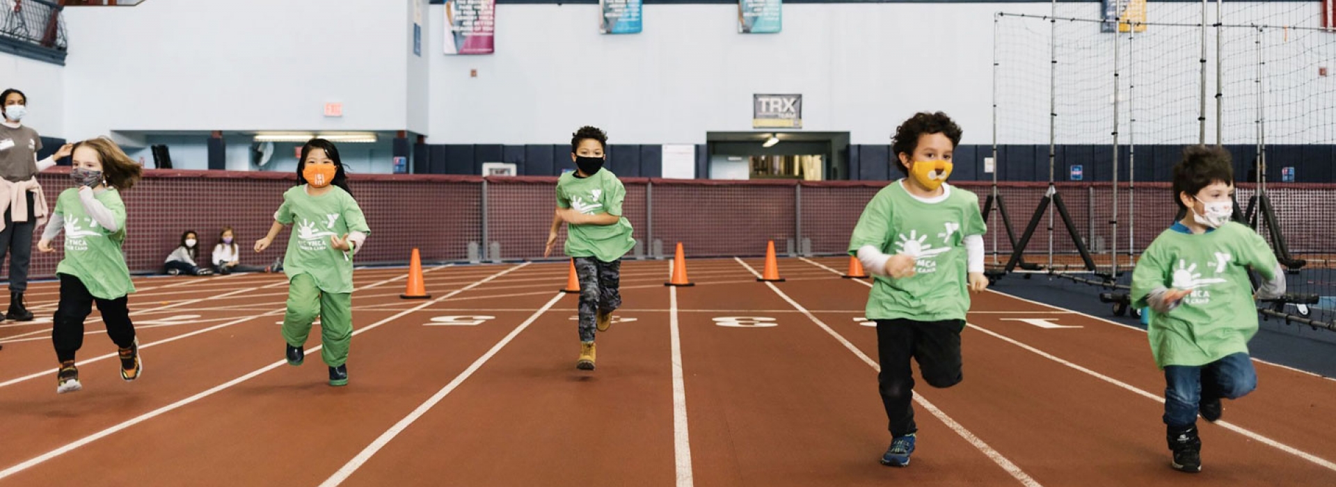 A group of kids in masks race on an indoor track during camp at the YMCA.